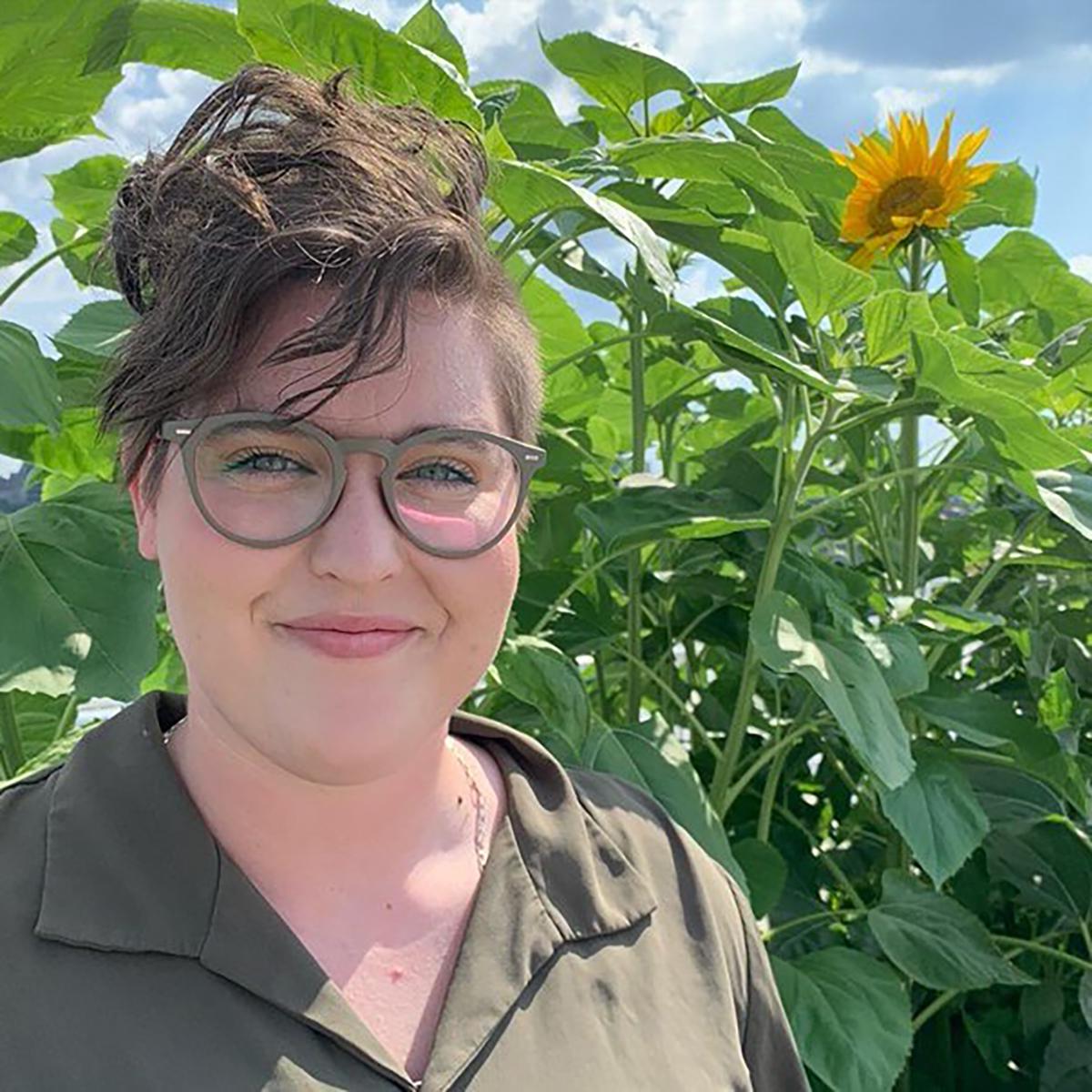 A smiling person in glasses stands in front of sunflowers on a bright sunny day